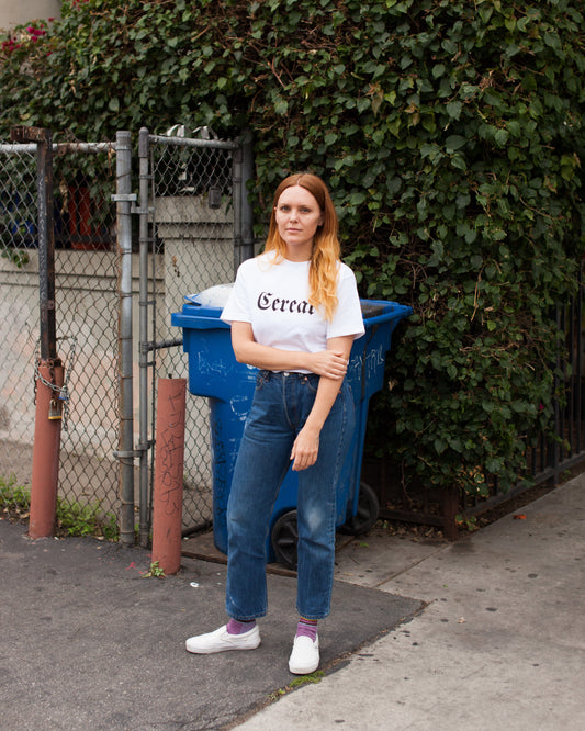 Person wearing a white t-shirt with 'Cereal' text, standing outdoors next to a blue trash bin.
