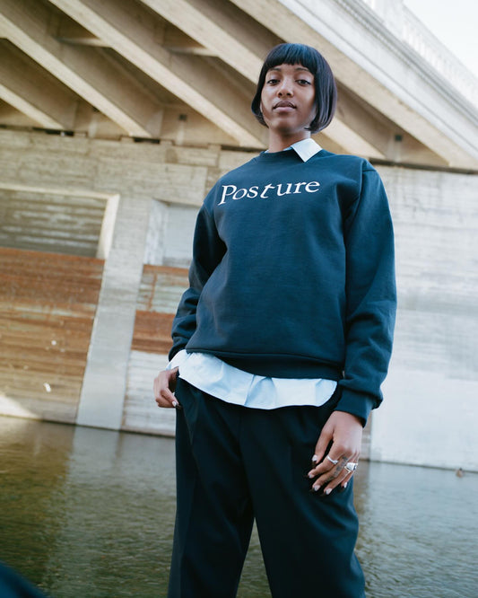 Person wearing a dark green sweatshirt with 'Posture' text, standing by a body of water.