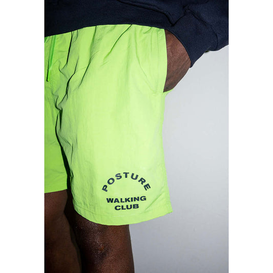 A person wearing neon green nylon team shorts with 3-pockets and a drawstring waist, featuring the text 'POSTURE WALKING CLUB' on the left leg.
