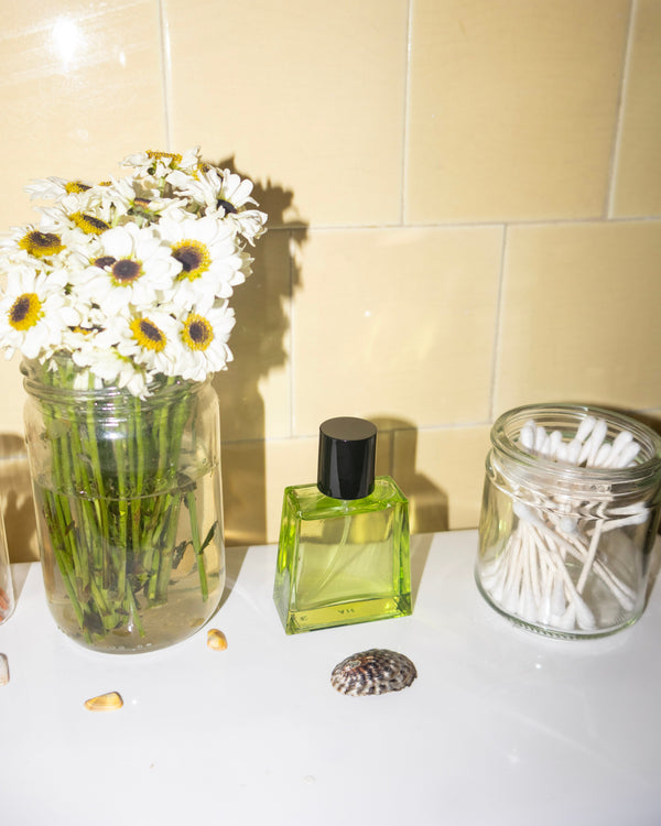 Vase with flowers, green bottle, and glass container on a white surface with beige tiled wall background