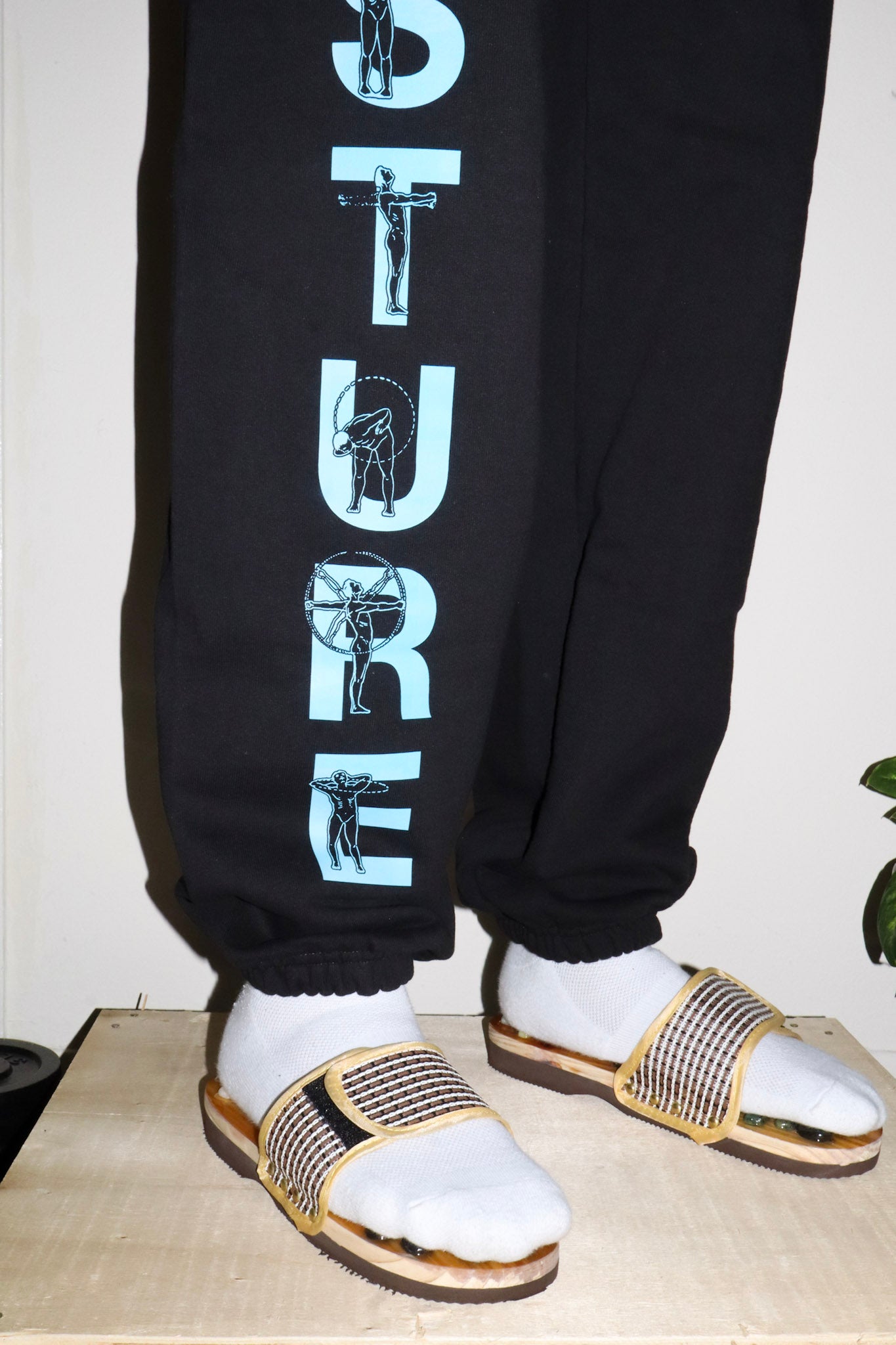 Person wearing black sweatpants with white text and white sandals on a light-colored floor.