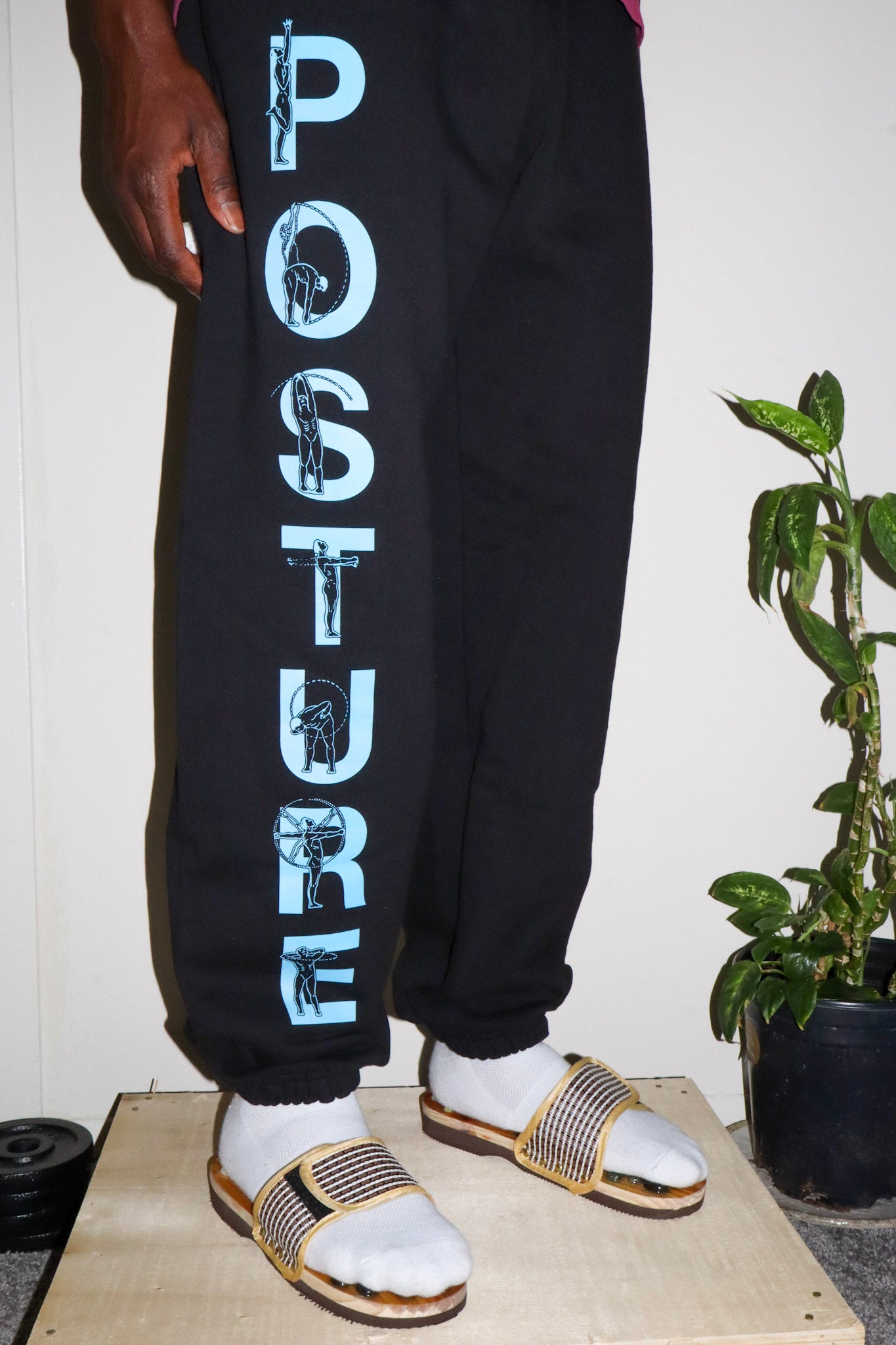 Person wearing black sweatpants with 'POSTURE' text, standing on a wooden platform with a plant in the background.