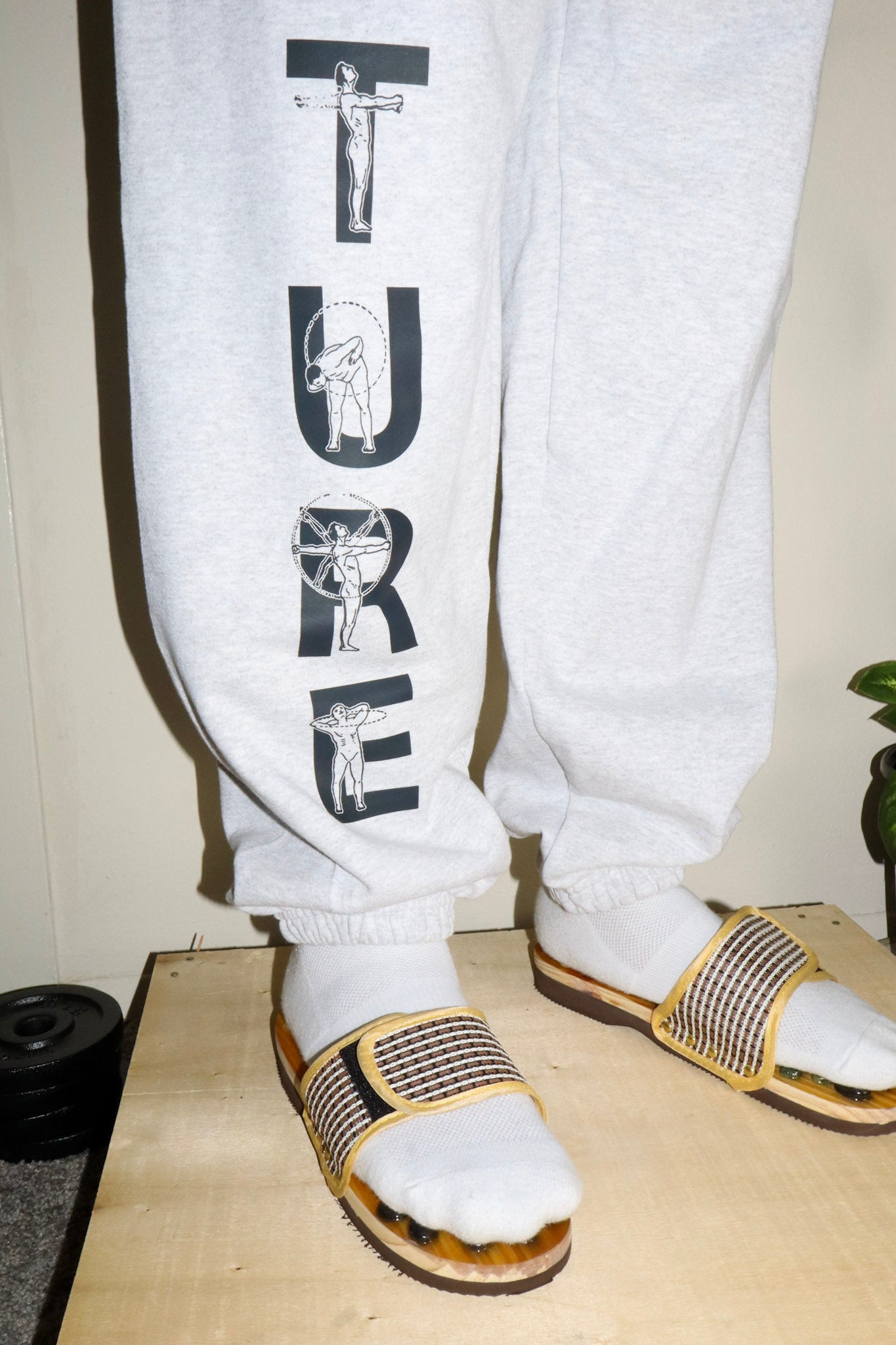 White sweatpants with black text and sandals on a wooden floor.