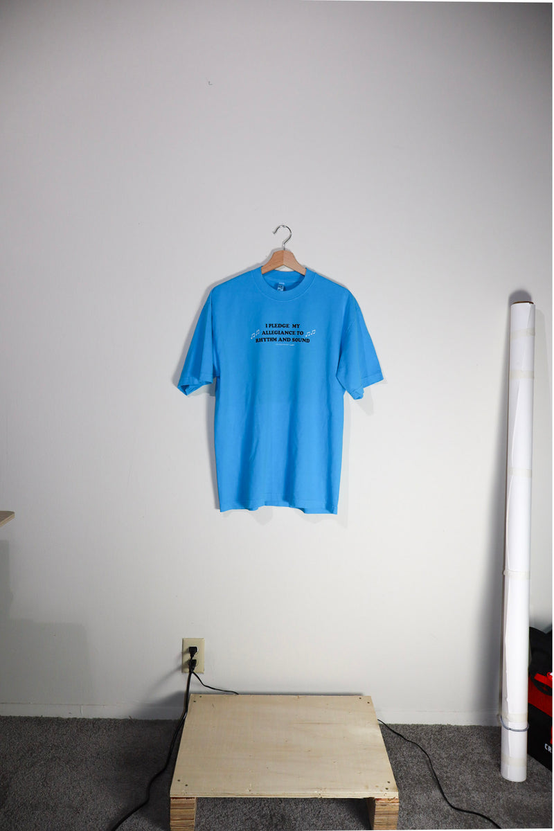 Blue t-shirt on a hanger against a white wall with a wooden block and white cylindrical object in the foreground.
