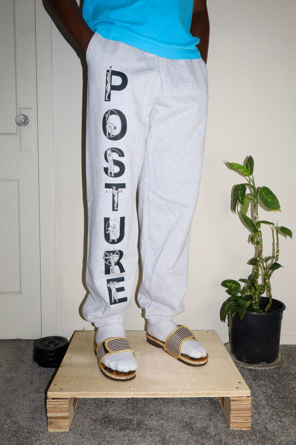 Person wearing light gray sweatpants with 'POSTURE' text, standing on a wooden platform with a plant in the background.