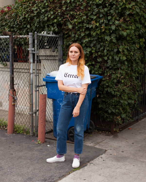 Person wearing a white t-shirt with 'Cereal' text, standing outdoors next to a blue trash bin.