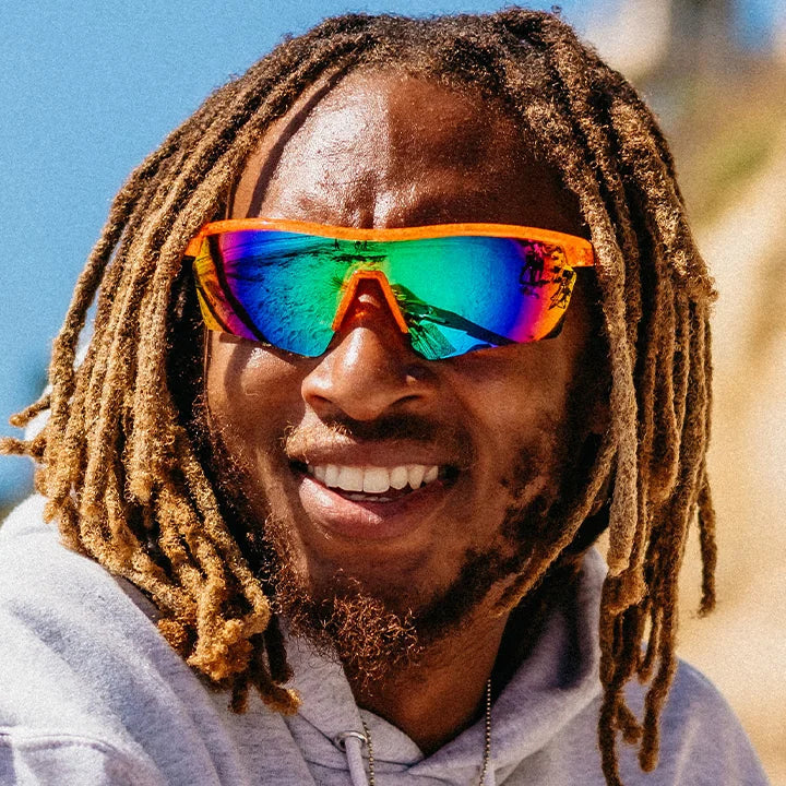 A smiling man with long dreadlocks wearing reflective shield-style sunglasses with a bright orange frame and mirrored lenses, photographed outdoors against a sandy hill and blue sky.