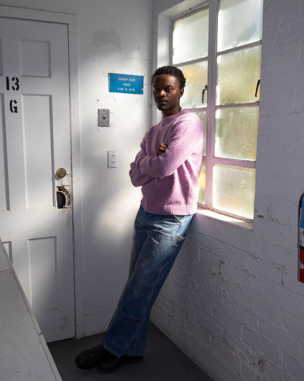 Person standing in a room with a door and window, wearing a pink sweater and blue jeans.