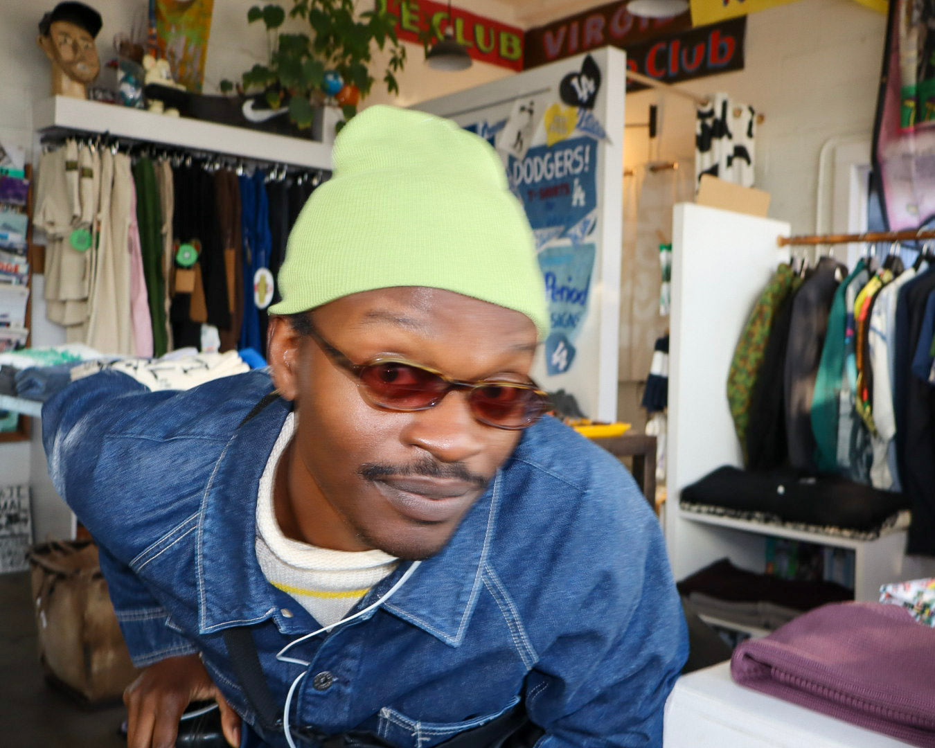 Person wearing a green beanie and sunglasses in a store setting with clothing racks and posters.