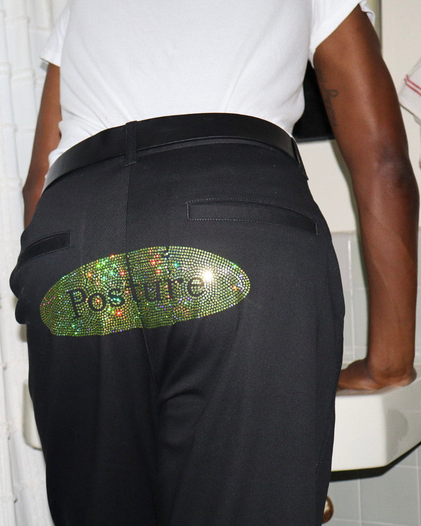 Black trousers with a large green rhinestone-embellished oval patch on the back that reads 'Posture' in black lettering, shown on a model wearing a white t-shirt.