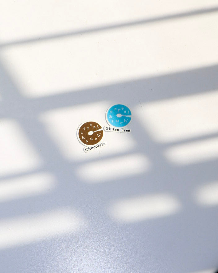 Two small oval stickers on a white surface, one brown labeled 'Chocolate' and one blue labeled 'Gluten-Free', with soft window shadow patterns across the scene.