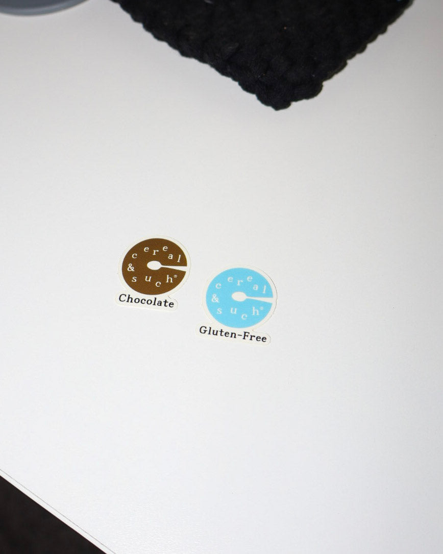 Two small oval stickers on a white surface, one brown labeled 'Chocolate' and one light blue labeled 'Gluten-Free', with the word 'Cereal' 