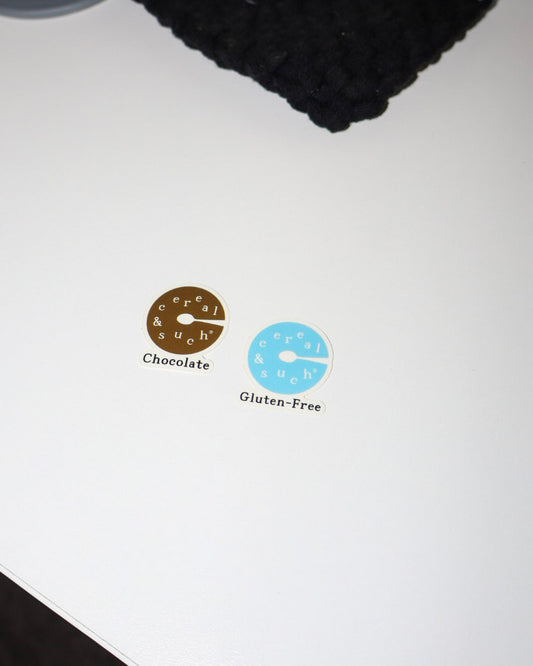 Two small oval stickers on a white surface, one brown labeled 'Chocolate' and one light blue labeled 'Gluten-Free', with the word 'Cereal' 