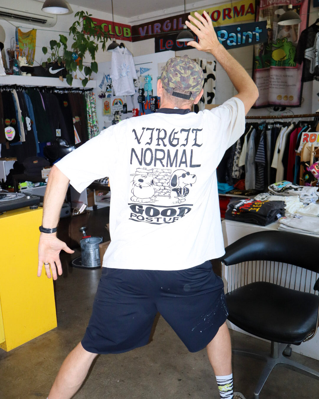 A man wearing a creme graphic t-shirt with a 'VIRGIL NORMAL GOOD POSTURE' cartoon print on the back, paired with dark shorts, posing inside a clothing store.