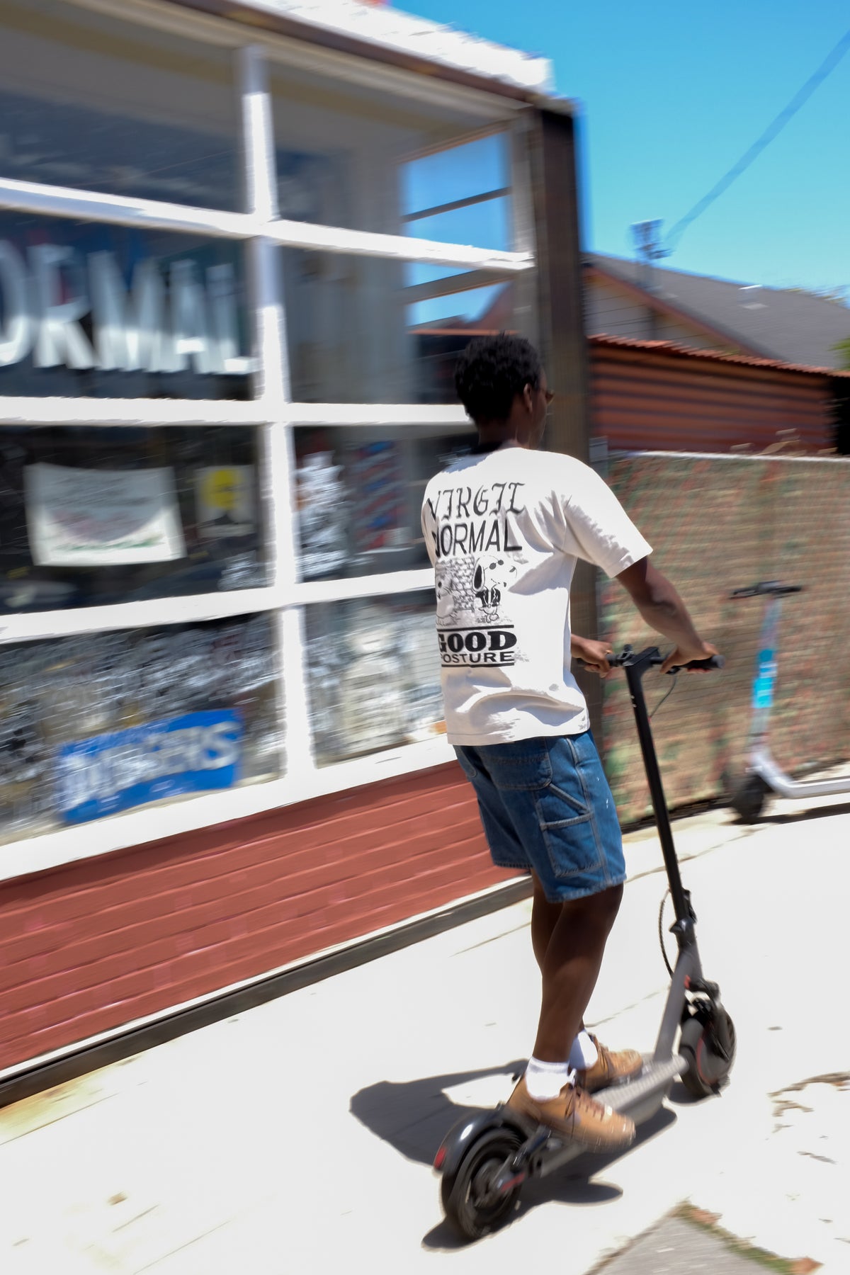 A man riding an electric scooter on a sidewalk next to a brick building, wearing a creme t-shirt with a black graphic print and blue denim shorts.
