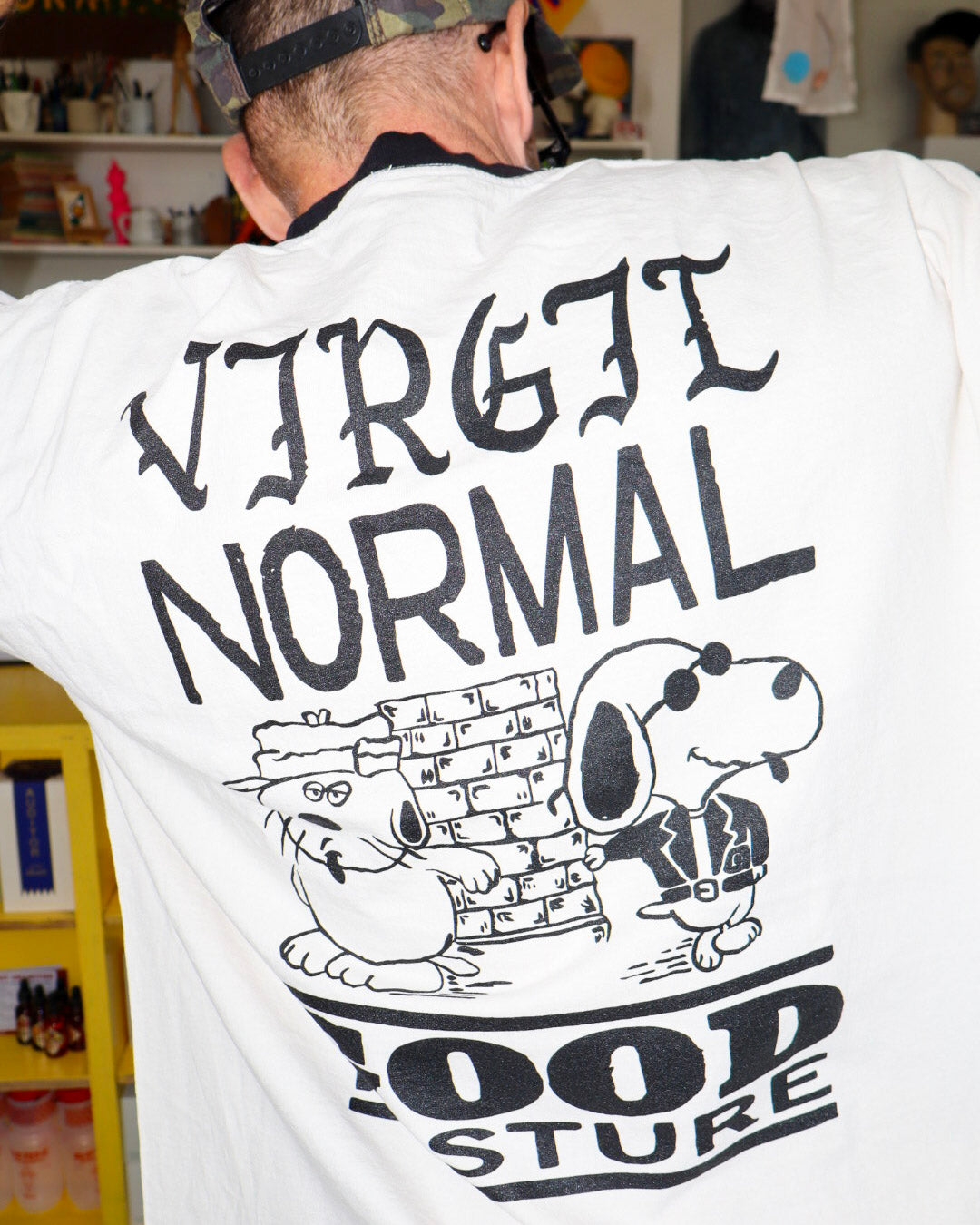 A person viewed from behind wearing a creme T-shirt with a large black graphic print featuring the text 'VIRGIL NORMAL' and 'GOOD POSTURE' with a cartoon dog and cat posed next to bricks.