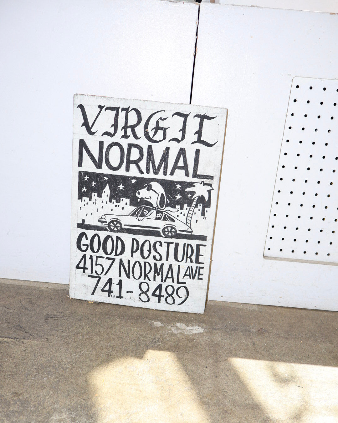 A rectangular white sign with black text reading 'VIRGIL NORMAL' and a black-and-white illustration of a person in a car, plus the text 'GOOD POSTURE 4157 NORMAL AVE 741-8489,' leaning against a white wall on a concrete floor.