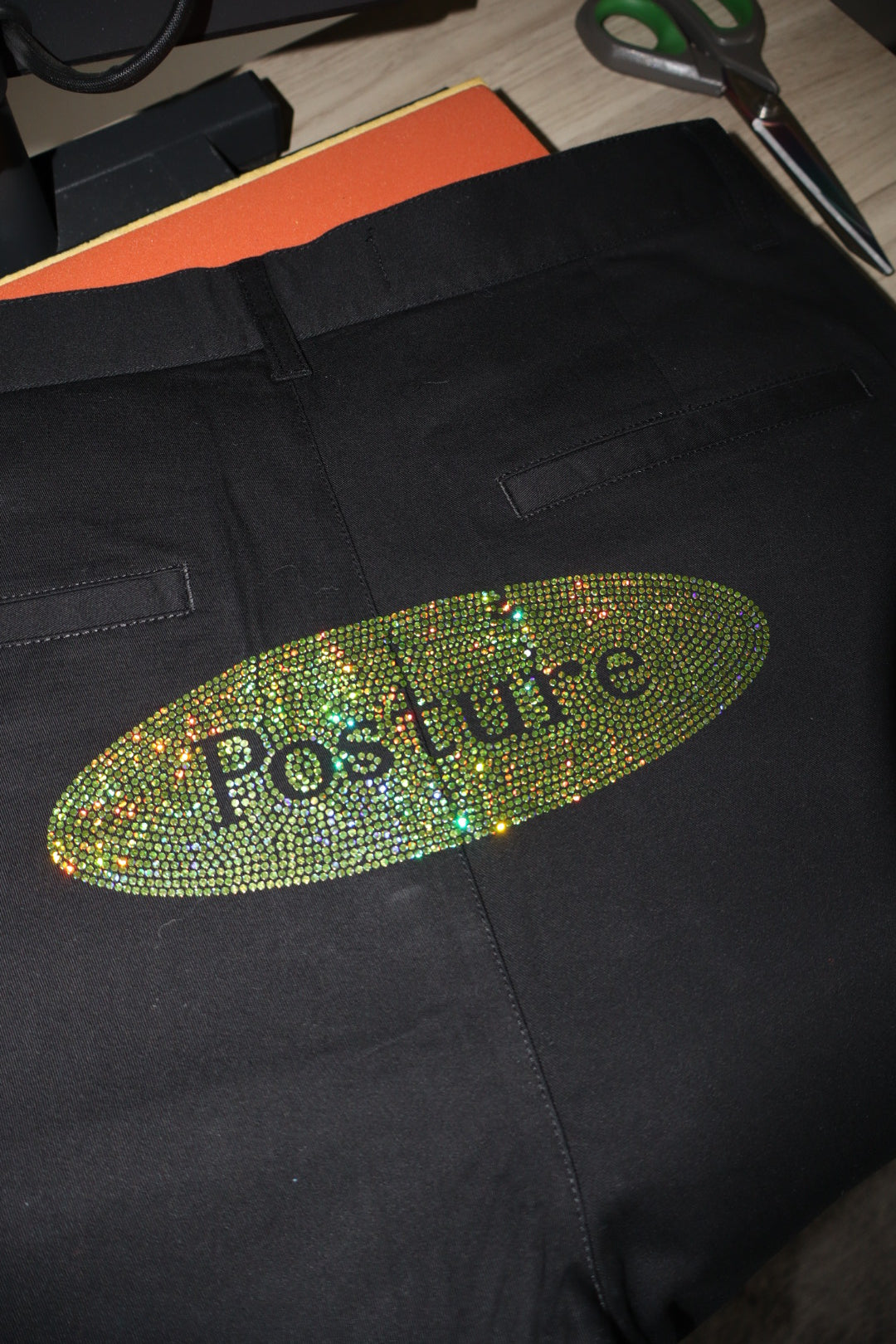 Black pants with an oval rhinestone-embellished patch on the back pocket, featuring the word 'Posture' in black script. The patch has a holographic, iridescent green and gold shimmer.
