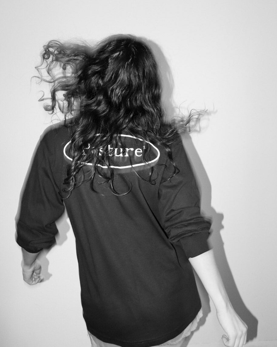 A person with long, dark, windswept hair is shown from behind wearing a black long-sleeve t-shirt with a white oval 'Future' logo printed across the upper back, set against a plain light gray wall.