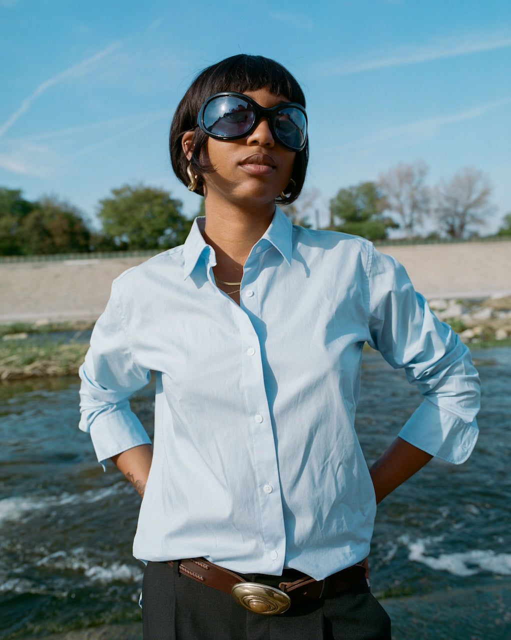 Person wearing a light blue shirt and sunglasses by a body of water with trees in the background.