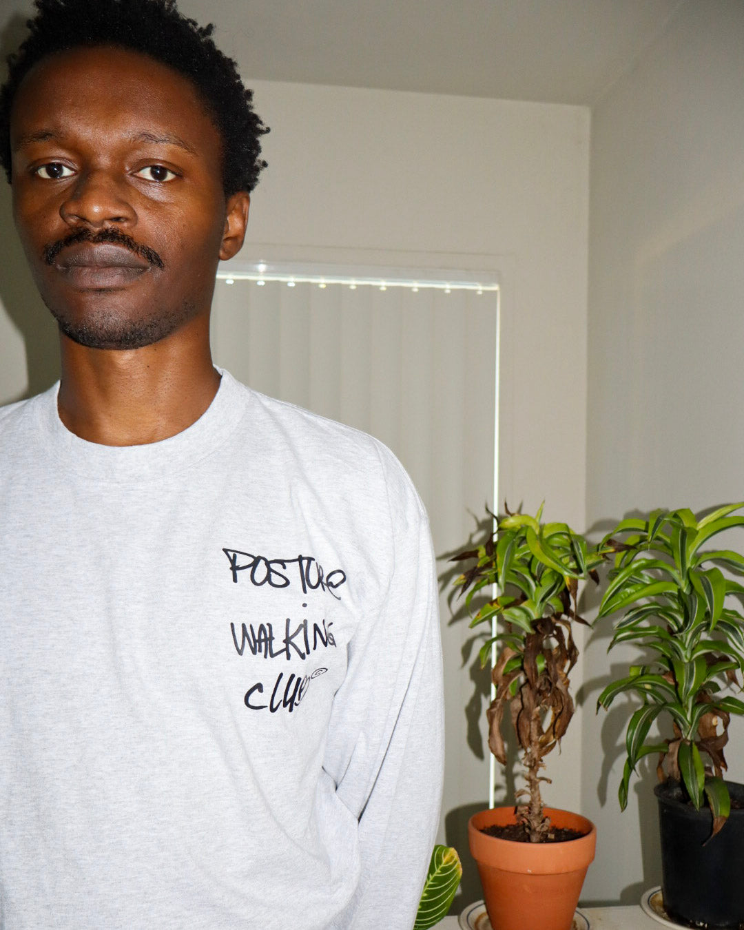 A man wearing a light gray crewneck sweatshirt with black handwritten text reading 'POSTURE WALKING CLUB' on the left chest, standing indoors next to potted plants.