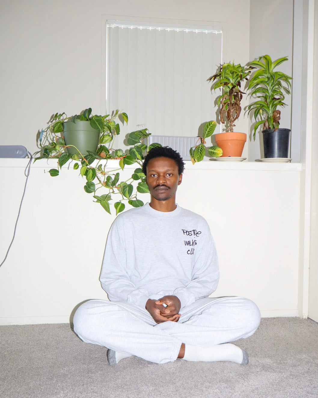 A man sits cross-legged on a gray carpet in a room with white walls and potted plants, wearing a light gray sweatshirt with black text and white sweatpants. The sweatshirt has the text 'Posture Walking Club' printed on the chest.