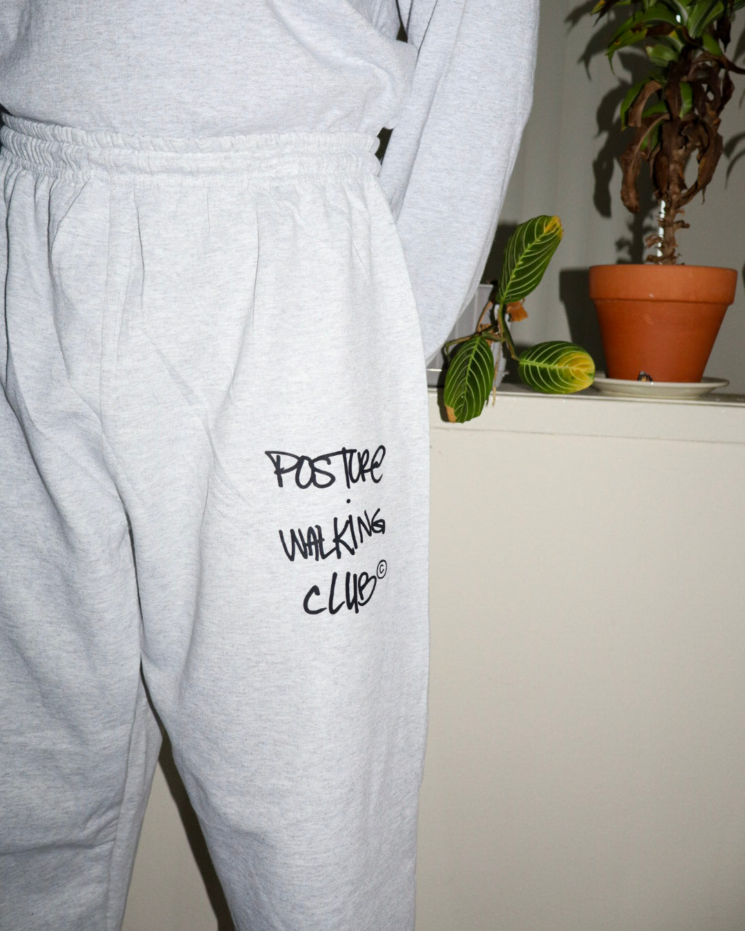 A person wearing light heather gray sweatpants with black handwritten text reading 'POSTURE WALKING CLUB' on the thigh, standing indoors near a potted plant.