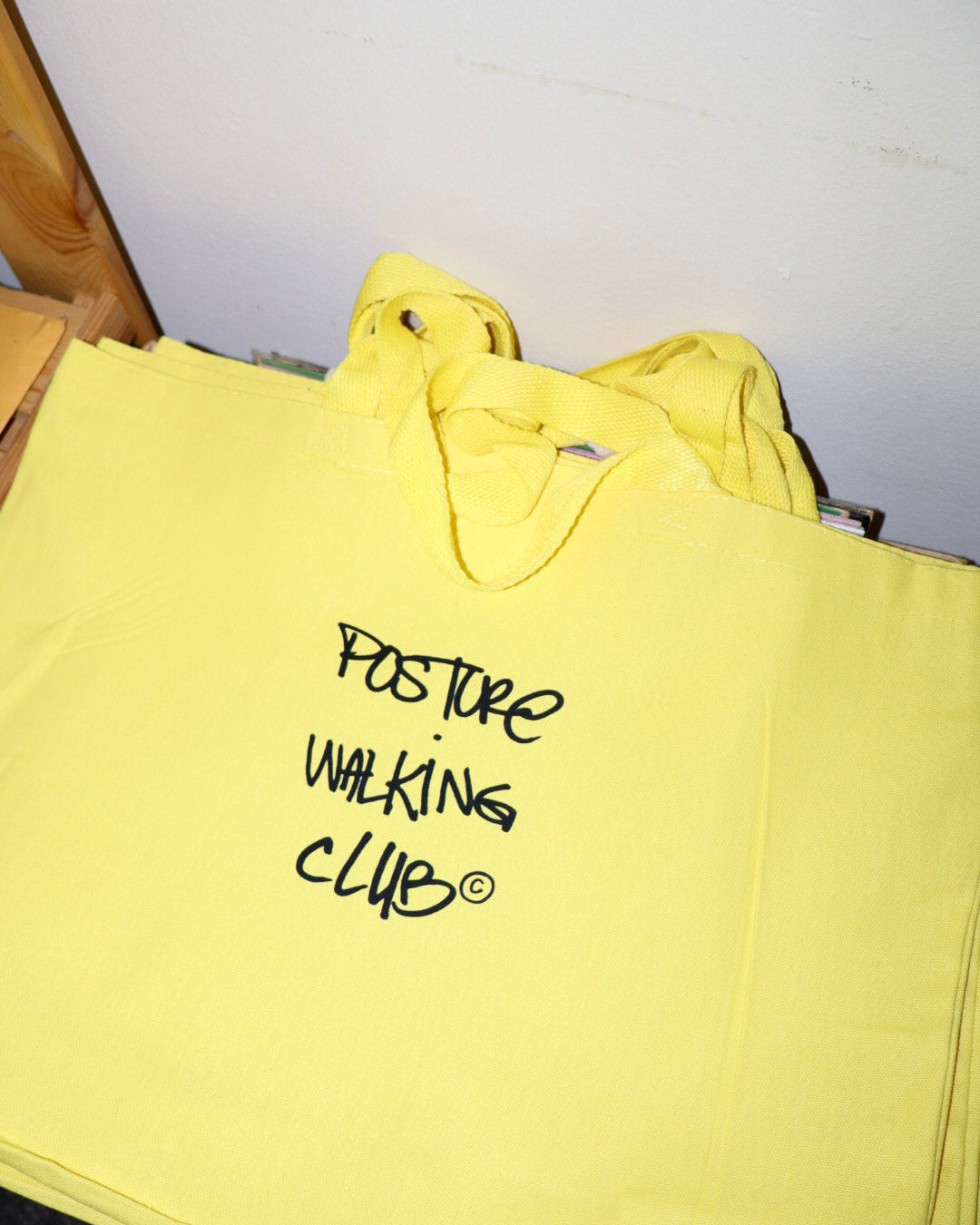 A bright yellow canvas tote bag with black handwritten text reading 'POSTURE WALKING CLUB' on the front, photographed indoors against a light wall.