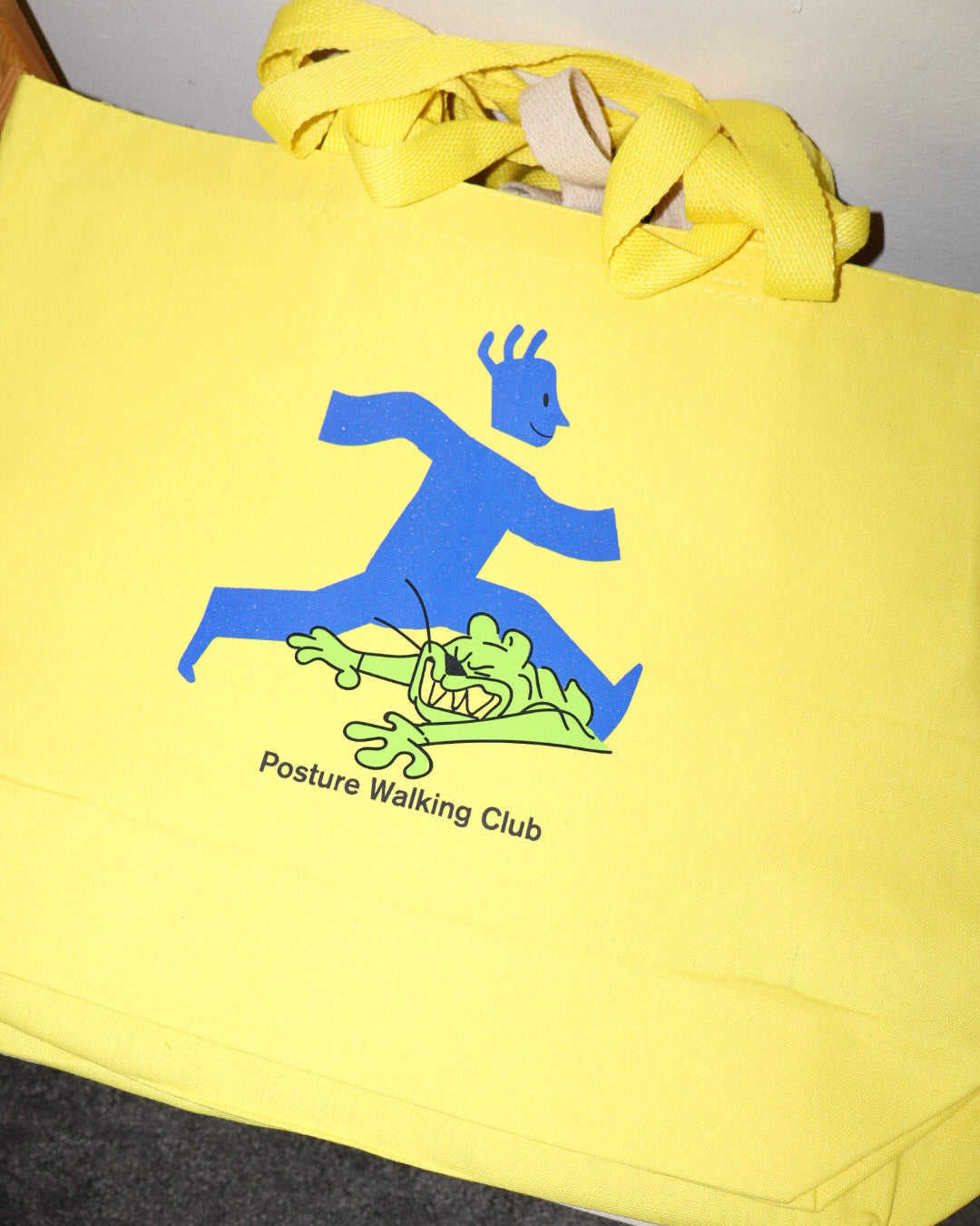 A bright yellow canvas tote bag with a blue and green graphic of a person walking over a cartoon creature, with the text 'Posture Walking Club' printed below.
