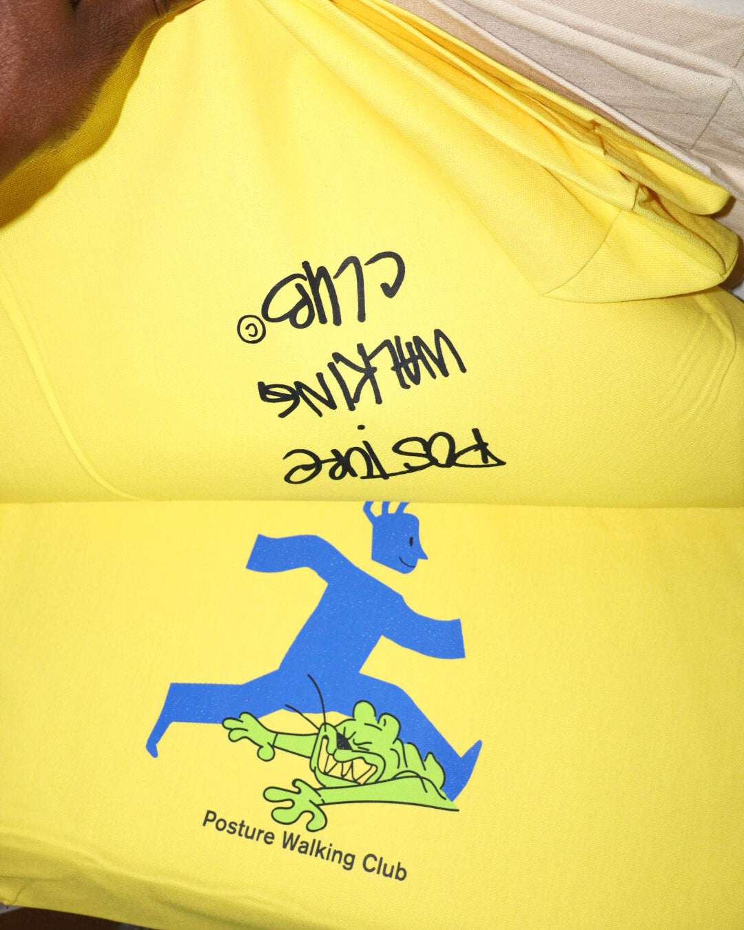 A bright yellow t-shirt featuring a graphic of a blue running figure pushing a green cartoon creature, with the text 'Posture Walking Club' printed below. The shirt is shown folded, with black handwritten text reading 'Posture Walking Club' written on the yellow fabric.