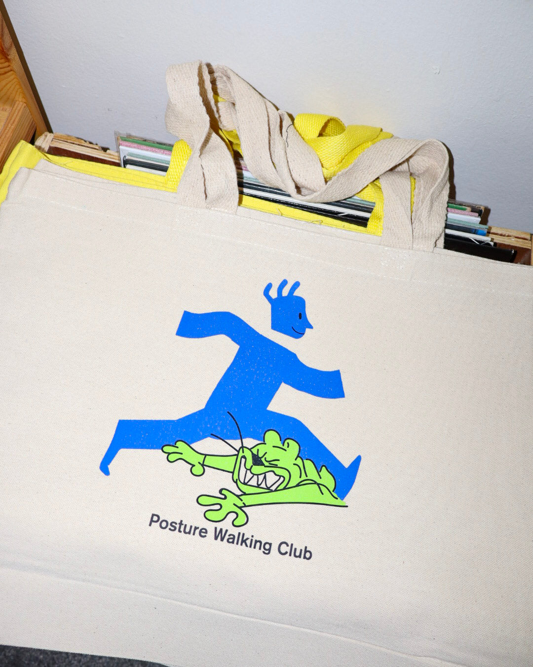 A cream canvas tote bag  featuring a blue walking figure pushing a green cartoon creature, with the text 'Posture Walking Club' printed below. The bag is shown resting on a shelf with books or files behind it.